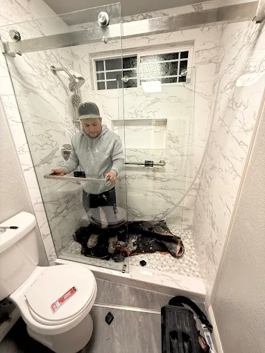 Modern marble tiled walk in shower with frameless glass enclosure being installed, featuring a built in niche, pebble stone floor, and window for natural light.